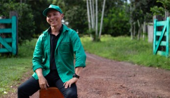 Homem sorrindo aluno do Senar e-Tec vestido de verde sentado em um banco em frente a um portão em uma fazenda
